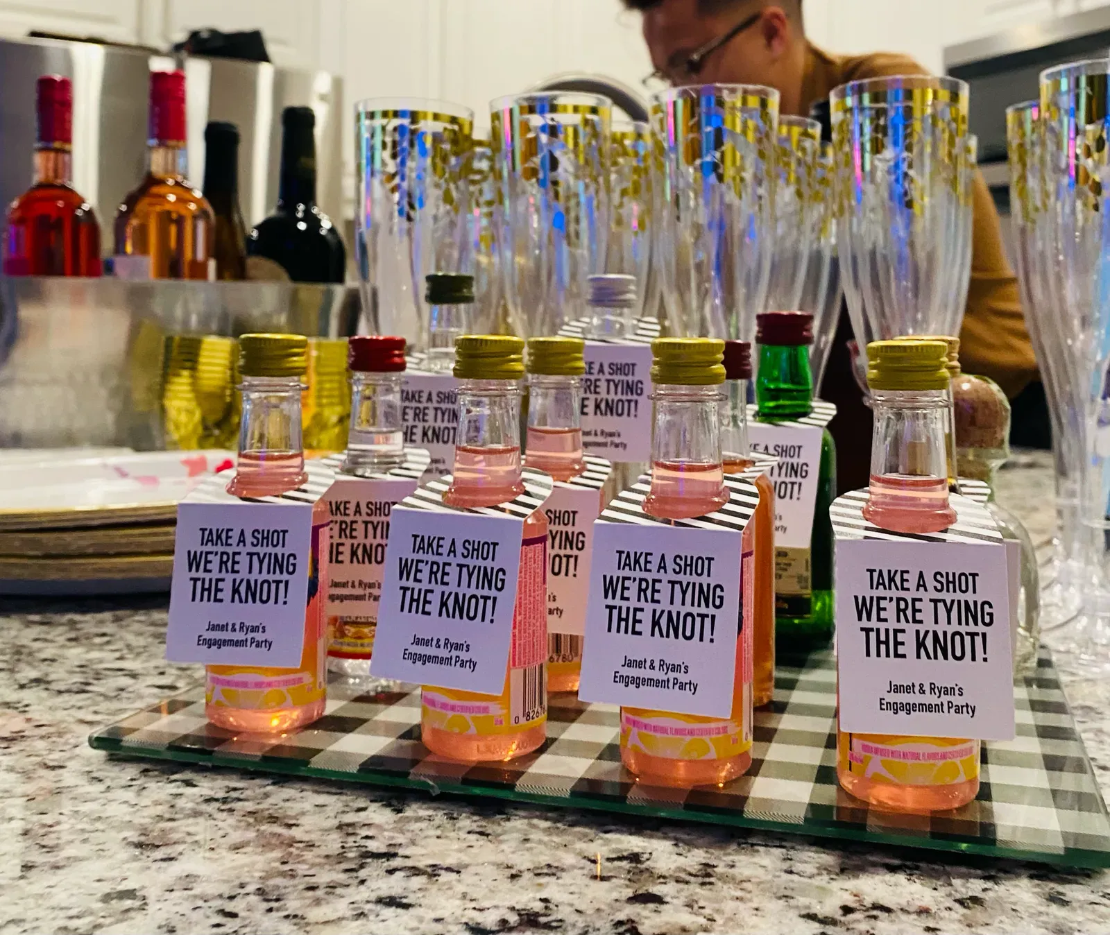 Rows of personalised mini drink favors with engagement party tags lined up on a kitchen counter